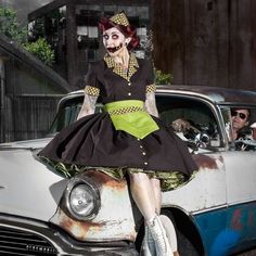 a woman in a dress is sitting on the hood of an old car with her face painted like a clown