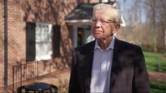 an older man standing in front of a red brick house wearing glasses and a blazer