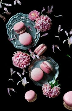 pink macaroons and flowers on glass plates with petals scattered around them in front of black background