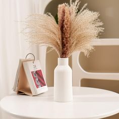 a white table with a vase and some dried grass in it next to a card holder