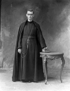 an old black and white photo of a man in a priest's robe standing next to a table