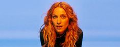 a woman with long red hair wearing a black shirt and looking at the camera while standing in front of a blue sky