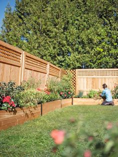 a woman sitting on the grass in front of a wooden fence and flower garden area