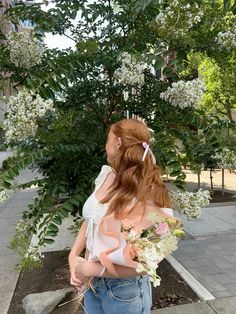 a woman with long red hair wearing a white top and blue jeans holding a bouquet of flowers