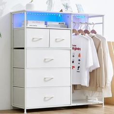 a white cabinet with drawers and shirts hanging on it's sides next to a plant