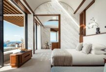 a bedroom with white walls and wood trimming on the ceiling, along with large windows overlooking the ocean