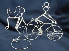 two wire figurines that are sitting on a blue cloth covered surface, one has a top hat and the other is holding a bicycle