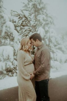 a pregnant couple standing next to each other in front of snow covered trees