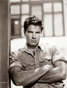 a black and white photo of a man with his arms crossed leaning against a pole