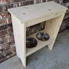 an unfinished wooden dog house with two bowls on the shelf and brick wall behind it