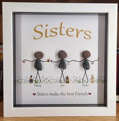 three rocks holding hands with the words sisters in gold lettering on them, sitting inside a white frame