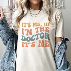 Stand out in style with our "It's Me Hi I'm the Doctor Its me" shirt. Designed for medical professionals, this shirt is the perfect conversation starter. Crafted from high-quality materials, it offers both comfort and durability for everyday wear. With its bold and quirky design, this shirt lets you proudly showcase your profession with a touch of humor. Whether you're in the hospital, clinic, or out and about, make a statement with the "It's Me Hi I'm the Doctor Its me" shirt. Discover comfort Vintage Cotton Tops With Funny Text, Retro Cotton Slogan Shirt, Retro Cotton Shirt With Funny Text, Hospital Clinic, Its Me, In The Hospital, Doctor Gifts, Med School, Quirky Design