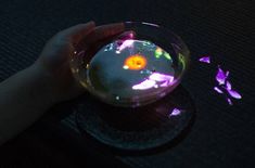 a hand is holding a glass bowl with flowers in it on a black tablecloth