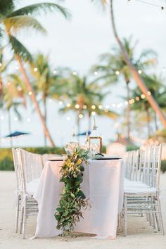 a table set up with white chairs and greenery