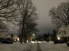 a snow covered street at night with cars parked on the side and houses in the background