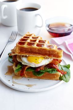 a waffle sandwich with bacon, egg and lettuce on a plate next to two cups of coffee