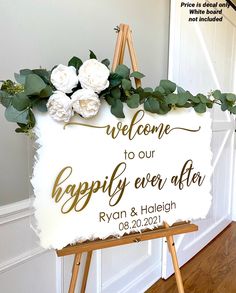 a welcome sign with flowers on it in front of a white wall and wooden easel