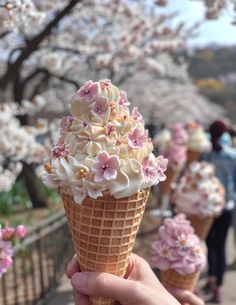 someone holding up an ice cream cone with pink and white flowers on it in front of cherry blossom trees