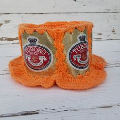 an orange crocheted cup holder with two beer labels on it, sitting on a white wooden surface