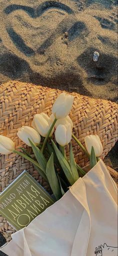 some white tulips are in a basket on the sand and there is a book