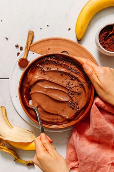 two hands holding a spoon over a bowl of chocolate pudding with bananas on the side