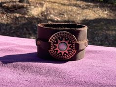 a brown leather cuff bracelet with an intricate design on the front and center, sitting on a purple surface