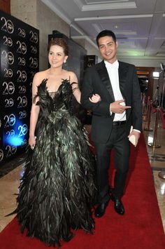 two people standing on a red carpet at an event wearing black and green dresses with feathers