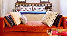 an orange couch with many pillows on it in front of two windows and a table