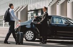 two men in suits are standing next to a black car and one man is holding a suitcase