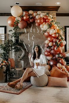 a woman sitting on the floor in front of a wall with balloons and greenery