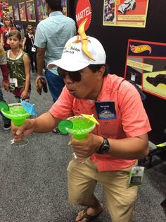 a man in an orange shirt and white hat is holding two green cups with yellow liquid