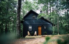 a black cabin in the woods with two chairs on the porch and one door open