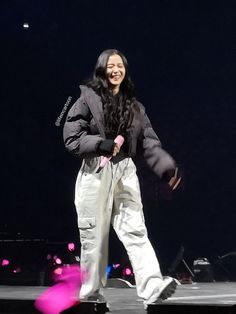 a woman standing on top of a stage wearing white pants and a black jacket with pink accents