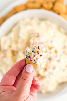 a hand holding a sprinkled donut over a bowl of dip and crackers