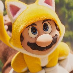 a close up of a stuffed animal wearing a yellow outfit and mustache with trees in the background