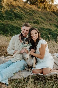 a man and woman sitting on a blanket with a dog in their lap smiling at the camera