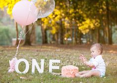 a baby sitting in the grass next to a pink cake and balloons with one on it