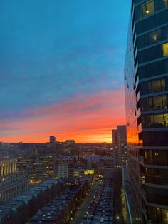 the sun is setting over a city with tall buildings and cars parked in front of it