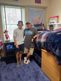 two men standing next to each other near a bed in a room with blue carpet