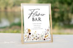 a flower bar sign sitting on top of a white tablecloth covered table next to a lake