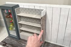 a person pointing at an open refrigerator with shelves in the front and behind it, on a wooden table