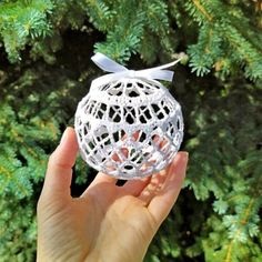 a hand holding a white ornament in front of a pine tree with a bow on it