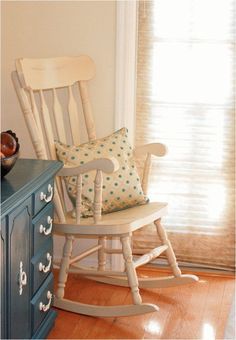 a rocking chair sitting in front of a window next to a blue dresser with a pillow on it