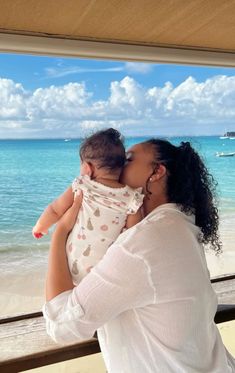 a woman holding a baby in her arms while looking out the window at the ocean