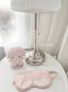 a pink sleeping mask next to a stuffed animal on a white table with a lamp in the background