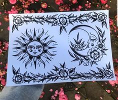 a person holding up a piece of paper with sun and moon designs on it in front of pink flowers