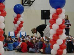 a group of children sitting on the floor in front of red, white and blue balloons