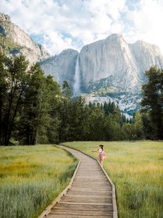 a woman standing on a wooden path in the middle of a field with mountains behind her