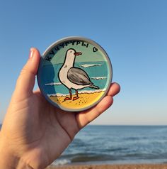 a hand holding up a button with a bird on the beach in front of the ocean