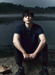 a young man sitting on top of a rock next to a body of water in front of a dark sky